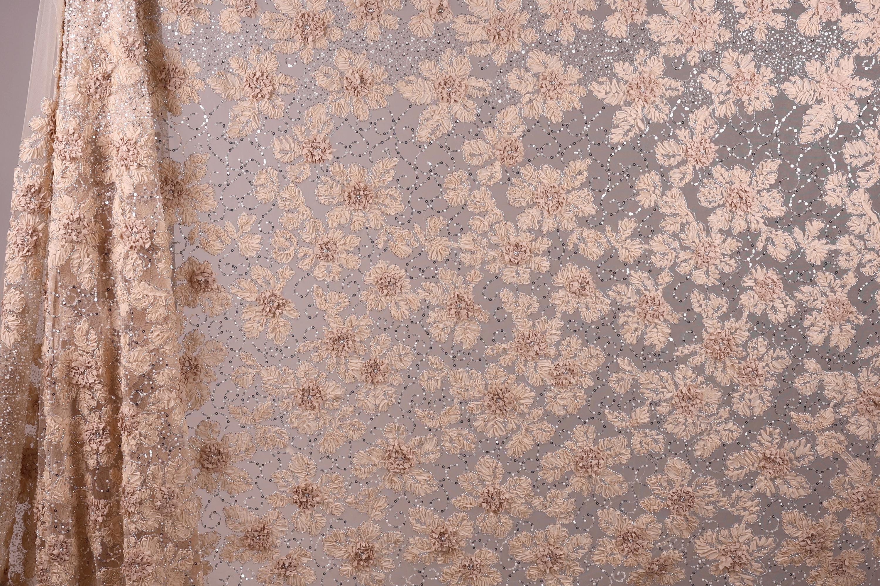 a close-up view of a fabric with a floral pattern, including small flowers and leaves. The fabric appears to be made of a light-colored material, possibly lace or a similar type of material. The pattern is intricate and detailed, with the flowers and leaves varying in size and shape. The image does not contain any text or other discernible objects, and the focus is solely on the floral pattern on the fabric.