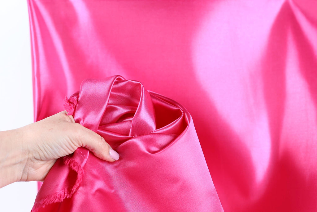 A hand is holding a piece of bright pink satin fabric, which appears to be a close-up view of the material.
