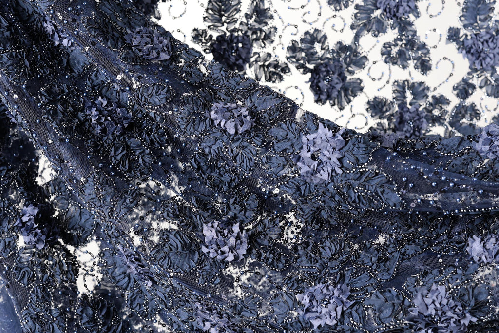 The image is a close-up view of a fabric with a floral pattern, featuring a mix of blue and black colors. The fabric appears to be made of lace or a similar material, with intricate designs and patterns created by the interlacing of the lace pieces. The overall appearance of the fabric is visually appealing and textured, with the contrasting colors and patterns creating a striking and eye-catching design.
