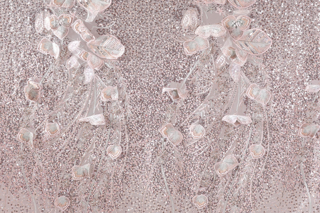 a close-up view of a fabric with a delicate, intricate design of pink and white flowers and leaves. The fabric appears to be made of sequin material, adding a sparkly, textured appearance. The flowers and leaves are arranged in a way that creates a sense of movement and depth, giving the fabric a three-dimensional effect. The image does not contain any text or other discernible objects, and the focus is solely on the floral design.