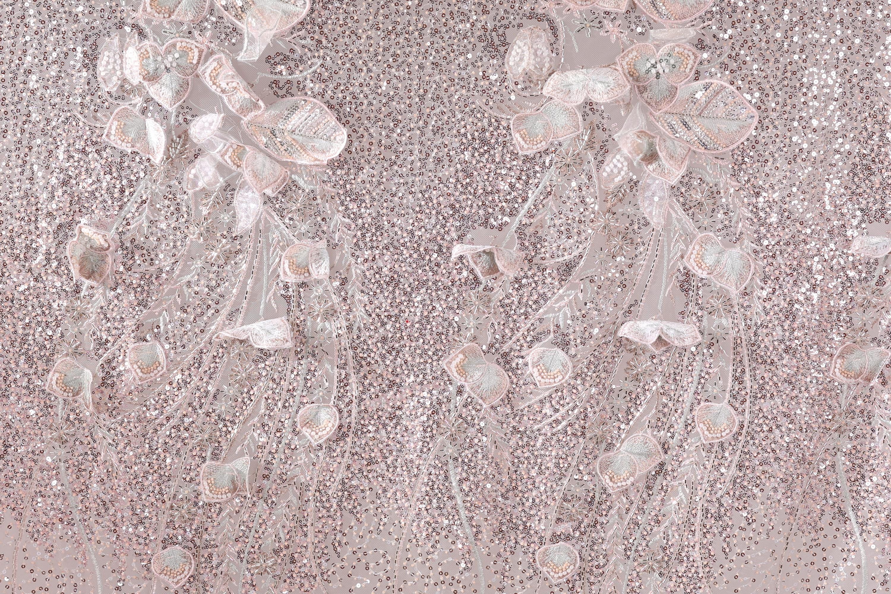 a close-up view of a fabric with a delicate, intricate design of pink and white flowers and leaves. The fabric appears to be made of sequin material, adding a sparkly, textured appearance. The flowers and leaves are arranged in a way that creates a sense of movement and depth, giving the fabric a three-dimensional effect. The image does not contain any text or other discernible objects, and the focus is solely on the floral design.