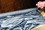 a roll of blue satin fabric with a vase of pink flowers placed next to it on a table.