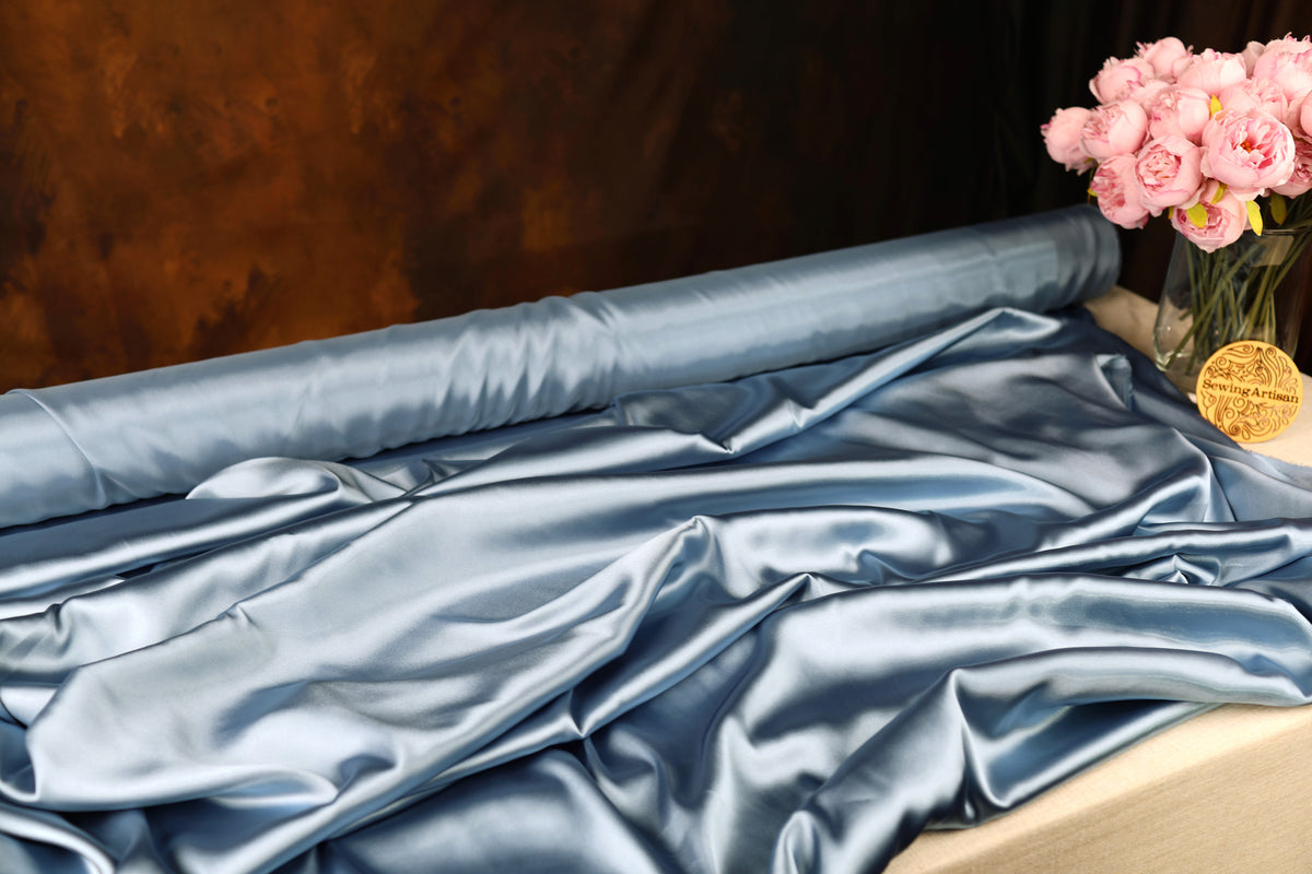 a roll of blue satin fabric with a vase of pink flowers placed next to it on a table.