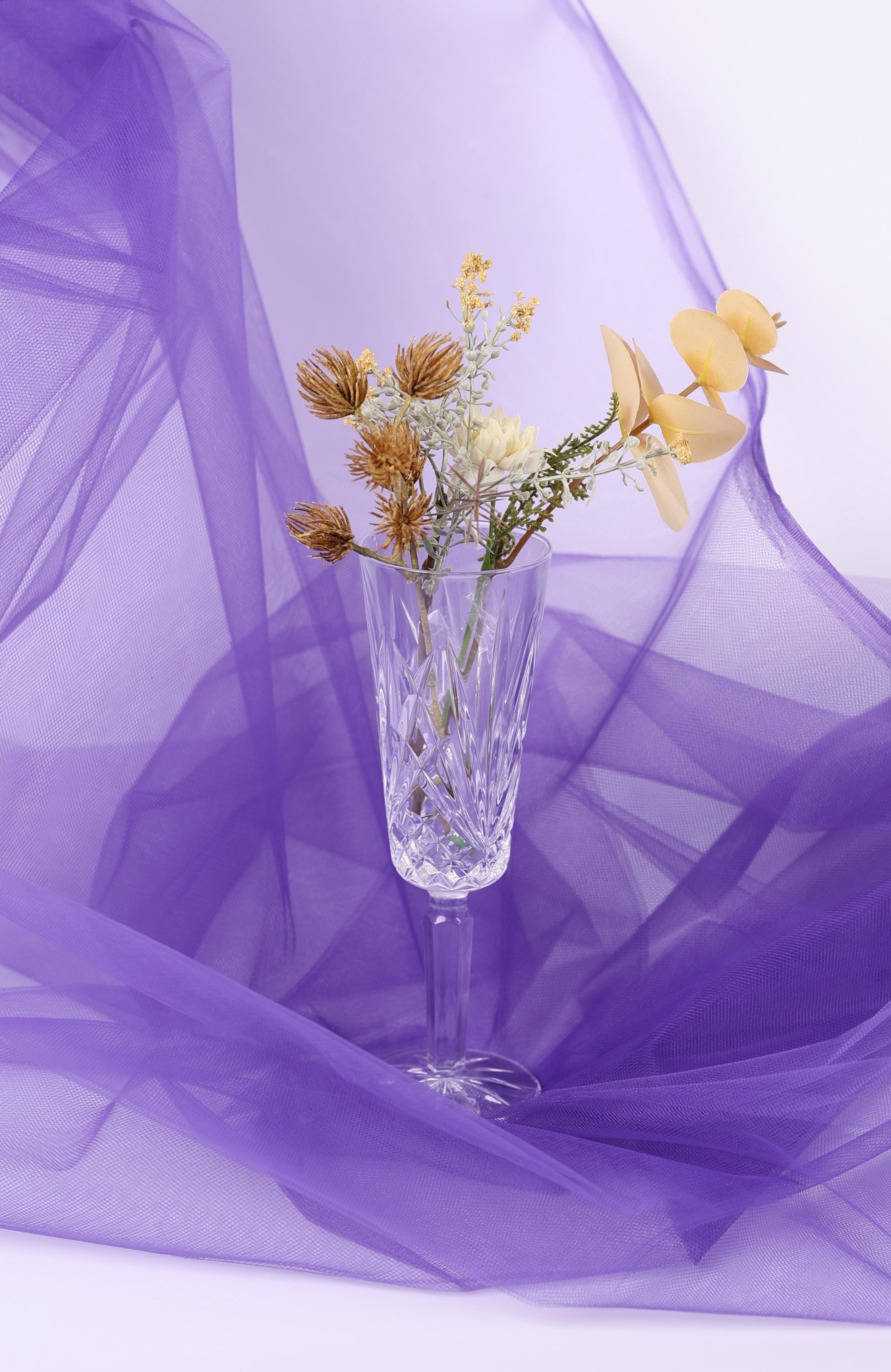 A clear glass vase filled with yellow flowers sits on a purple fabric background.