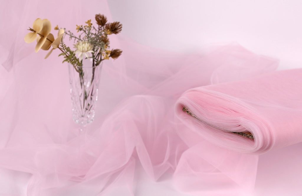 A clear glass vase filled with dried flowers sits on a pink fabric, with a folded piece of the fabric next to it.
