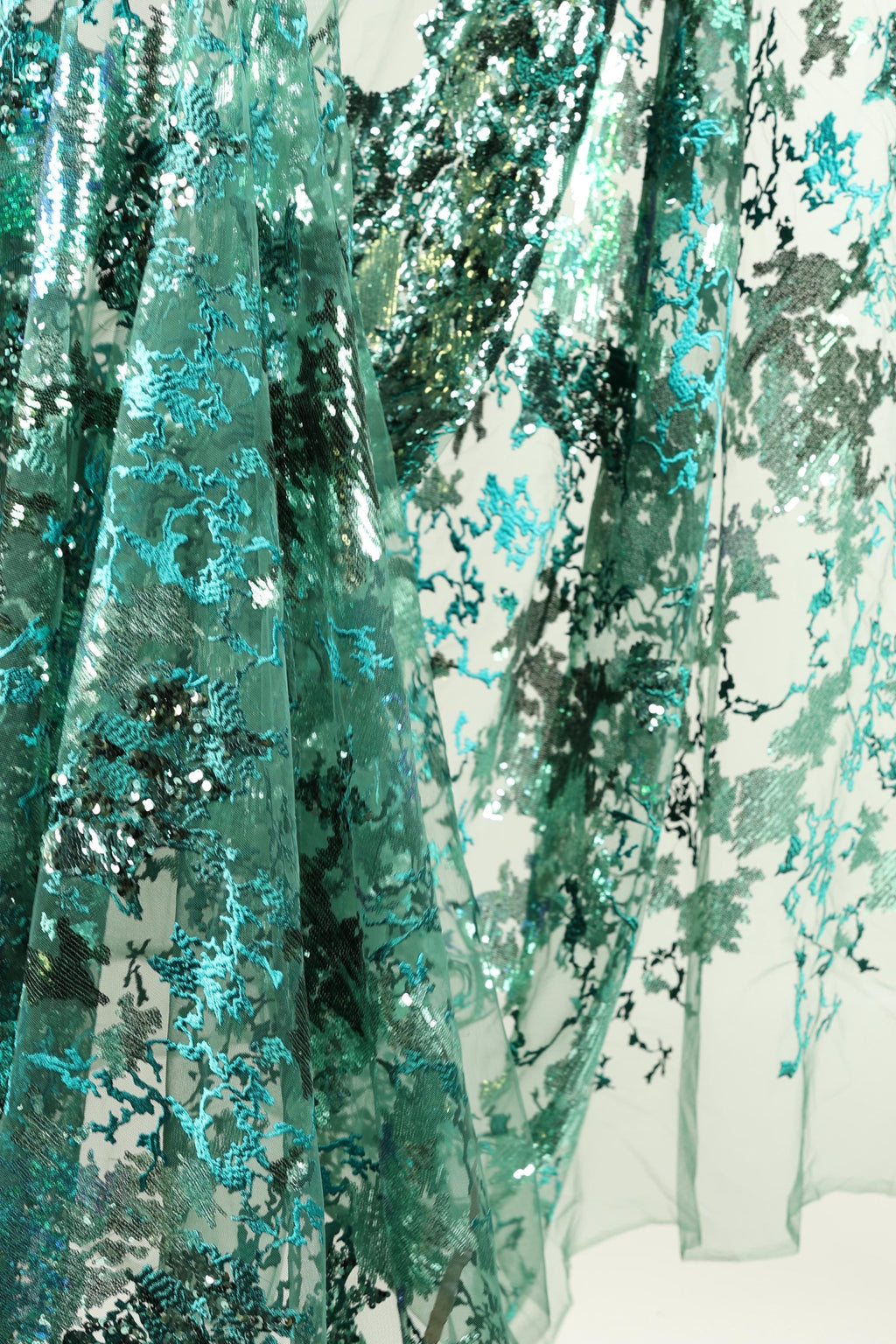 The image is a close-up of a fabric with a floral pattern, featuring a mix of green and blue colors. The fabric appears to be made of sheer material, with a textured and intricate design.