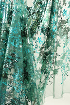 The image is a close-up of a fabric with a floral pattern, featuring a mix of green and blue colors. The fabric appears to be made of sheer material, with a textured and intricate design.
