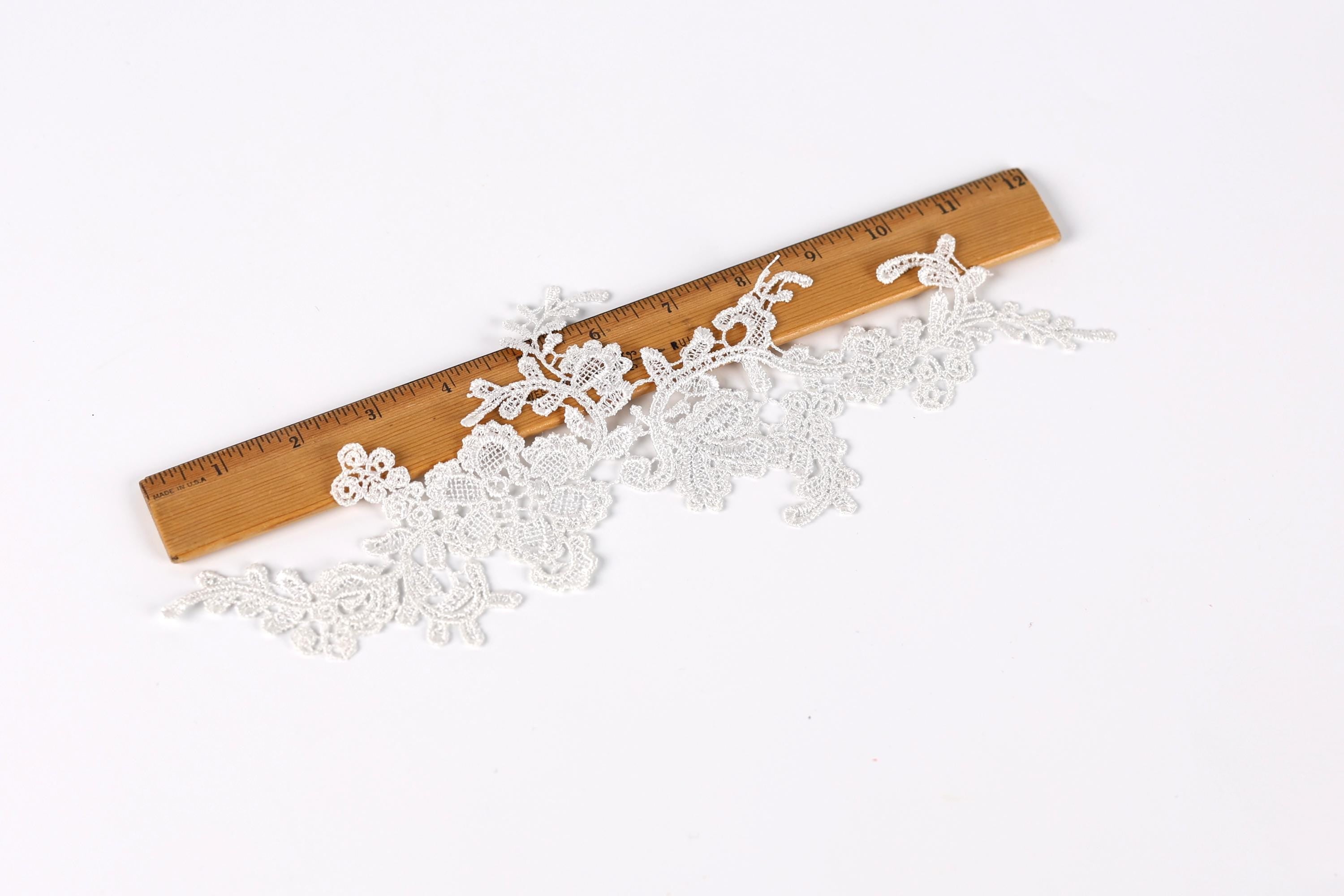 a piece of white lace with intricate floral patterns lying on a wooden ruler, which is placed on a white background.