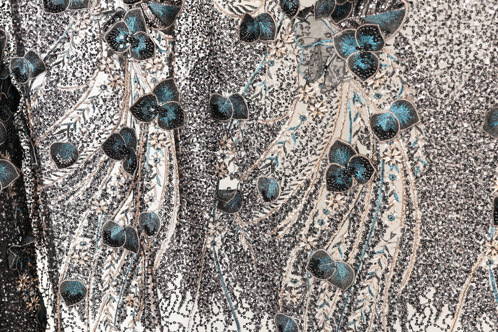a close-up view of a fabric with a floral pattern, including blue and green leaves and flowers. The fabric appears to be made of sequin material, creating a textured and visually striking appearance.