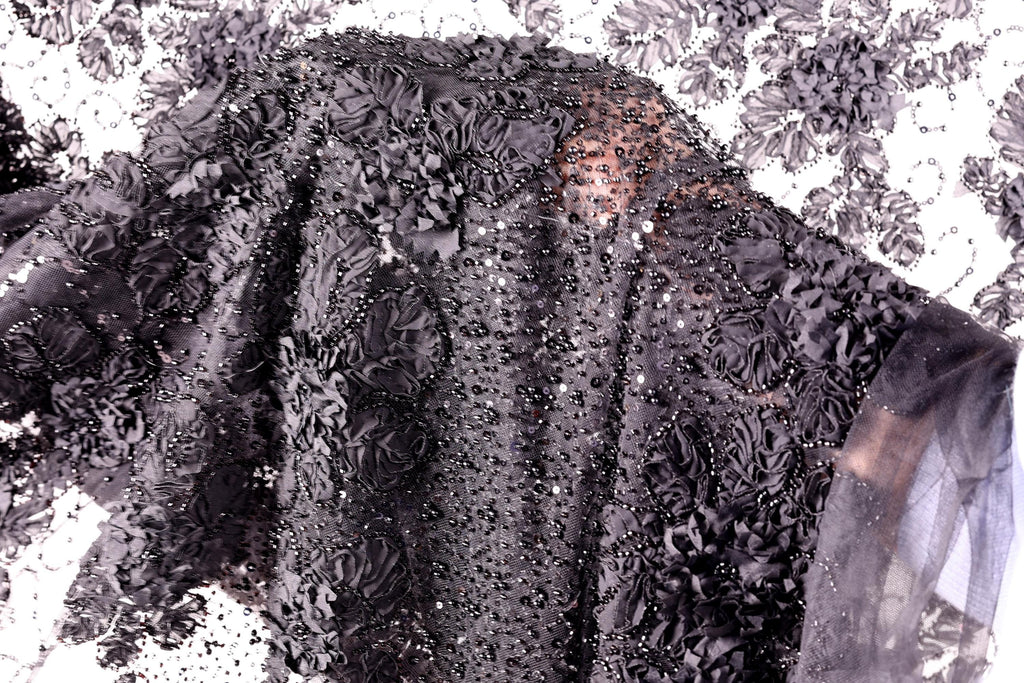 a close-up view of a black fabric with a floral pattern, including black flowers and sequins. The fabric appears to be a black dress or garment with a detailed, intricate design.