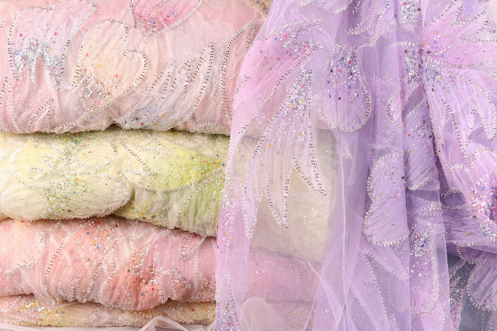 a close-up view of a stack of fabric swatches in various pastel colors, including pink, yellow, and purple. The fabric appears to be made of a sheer, lace-like material, with intricate beading and embroidery visible on the surface.