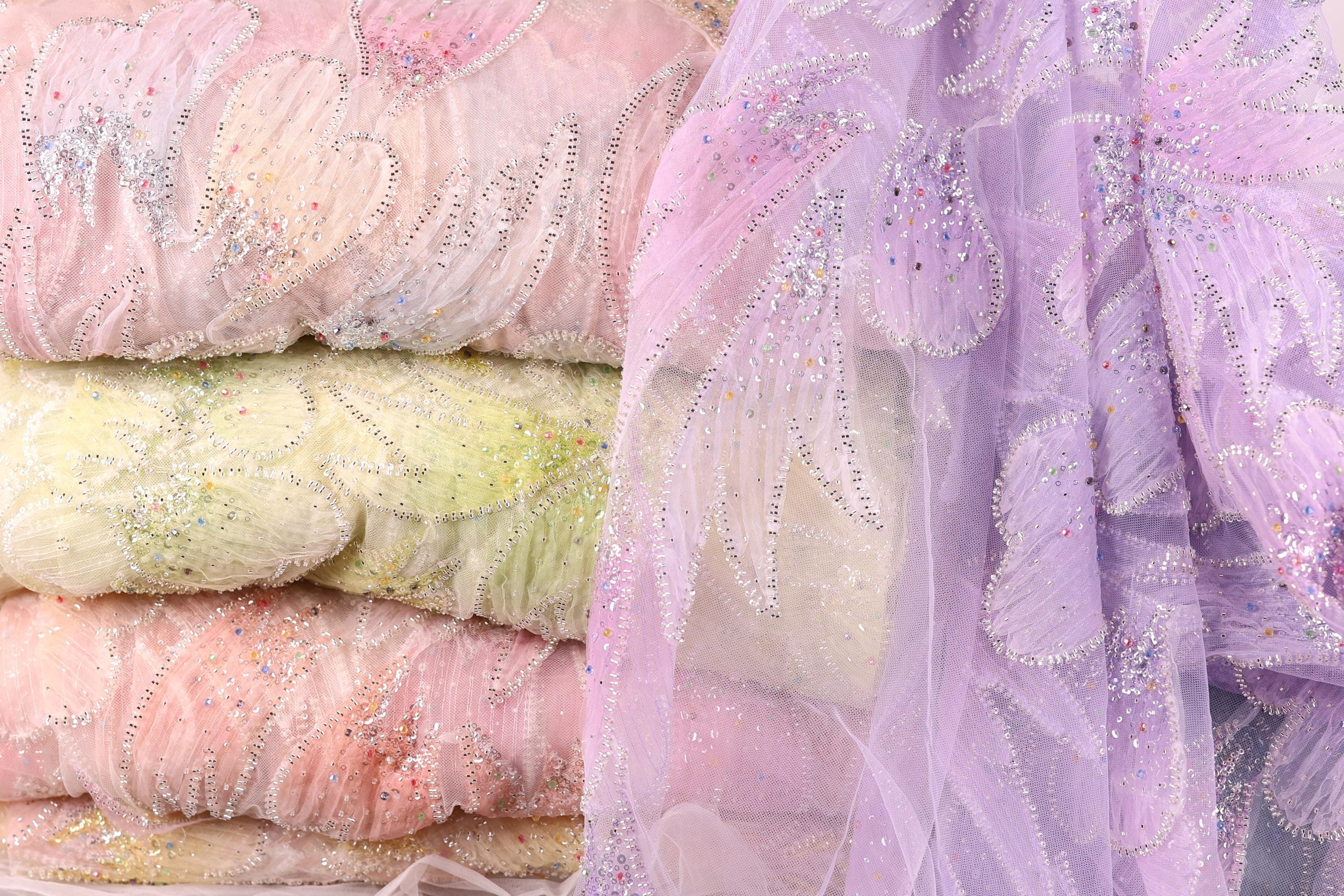 a close-up view of a stack of fabric swatches in various pastel colors, including pink, yellow, and purple. The fabric appears to be made of a sheer, lace-like material, with intricate beading and embroidery visible on the surface.