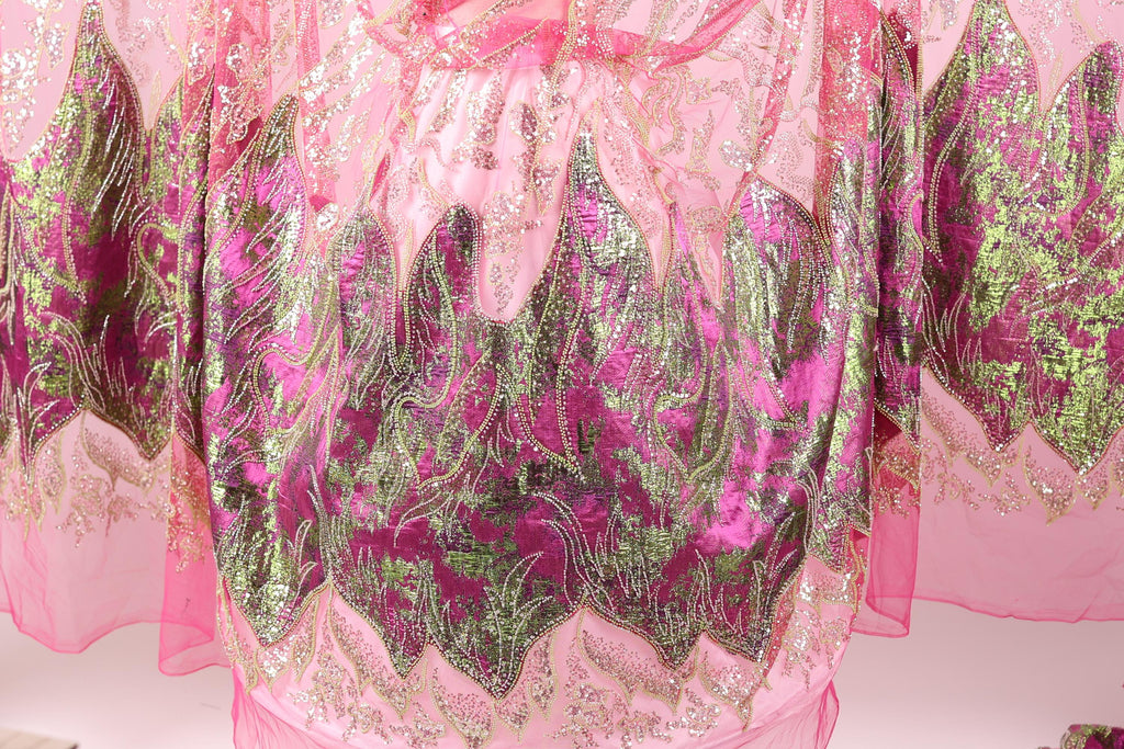a close-up view of a pink and green fabric with intricate, sparkling designs. The fabric appears to be a sheer or sheer-like material, with a floral pattern in shades of pink and green. The design elements are detailed and visually striking, creating a vibrant and eye-catching appearance.