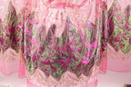 a close-up view of a pink and green fabric with intricate, sparkling designs. The fabric appears to be a sheer or sheer-like material, with a floral pattern in shades of pink and green. The design elements are detailed and visually striking, creating a vibrant and eye-catching appearance.