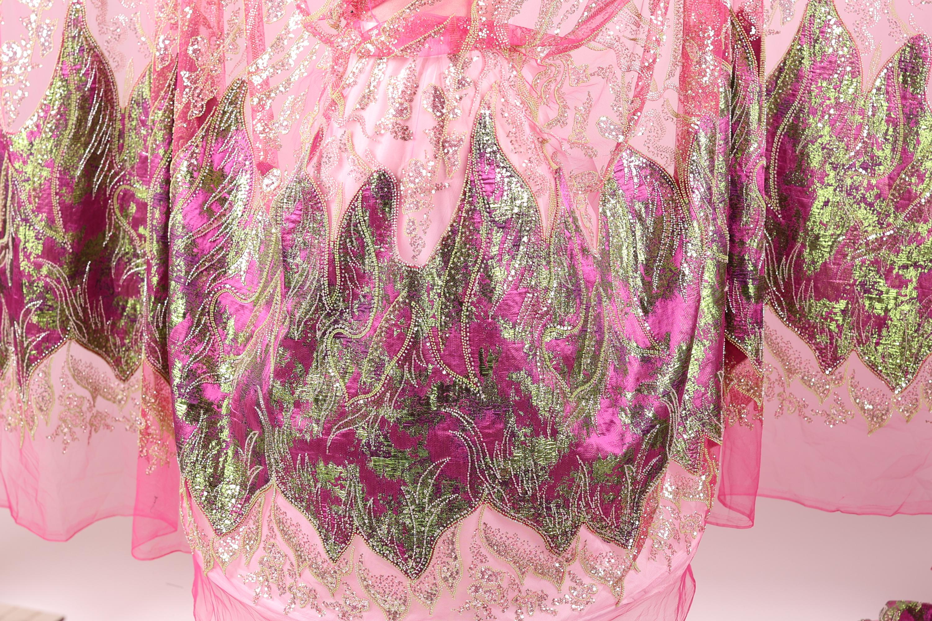 a close-up view of a pink and green fabric with intricate, sparkling designs. The fabric appears to be a sheer or sheer-like material, with a floral pattern in shades of pink and green. The design elements are detailed and visually striking, creating a vibrant and eye-catching appearance.