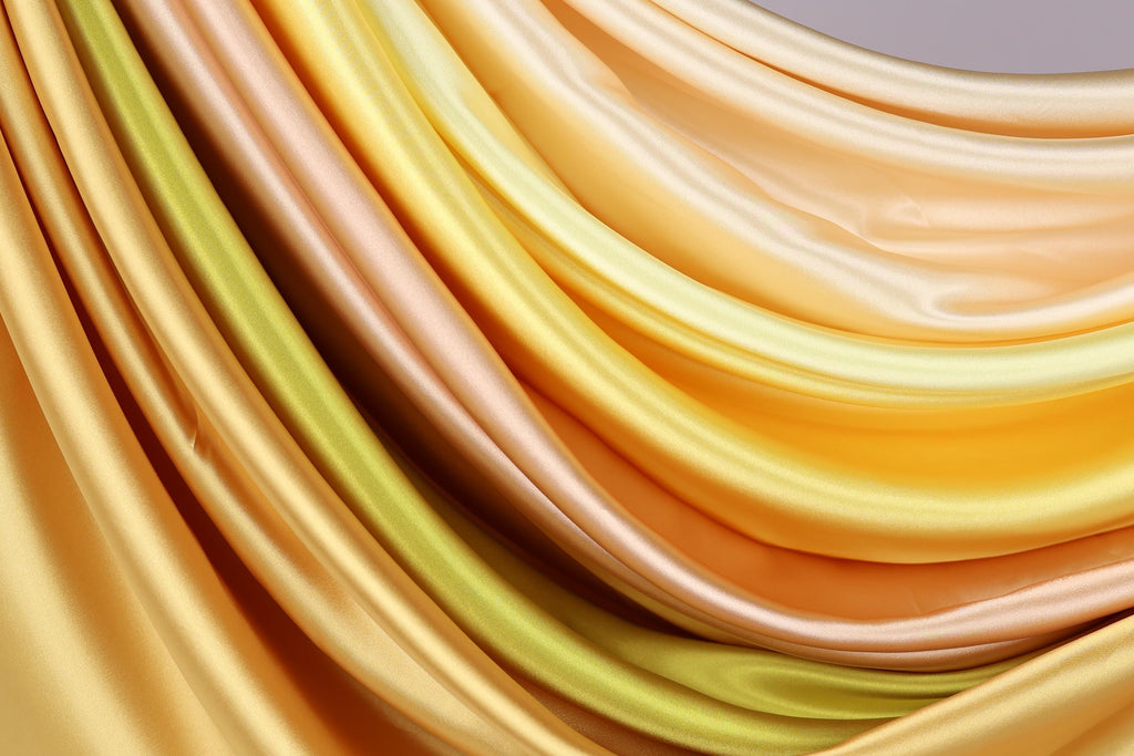 a close-up view of a fabric with a vibrant, multicolored pattern. The fabric appears to be made of silk or satin, with a smooth and shiny texture. The colors of the fabric are a mix of yellow, orange, and green, creating a visually striking and dynamic composition.
