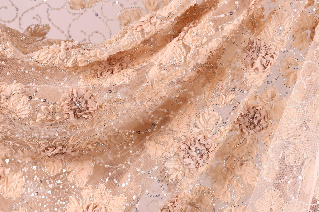 a close-up view of a delicate, intricately embroidered fabric with a floral pattern. The fabric appears to be made of a light-colored material, possibly lace or chiffon, with a floral design that includes various shades of pink and beige flowers and leaves. The fabric is adorned with small, sparkling embellishments, adding a touch of elegance and sophistication to the overall appearance.