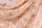 a close-up view of a delicate, intricately embroidered fabric with a floral pattern. The fabric appears to be made of a light-colored material, possibly lace or chiffon, with a floral design that includes various shades of pink and beige flowers and leaves. The fabric is adorned with small, sparkling embellishments, adding a touch of elegance and sophistication to the overall appearance.