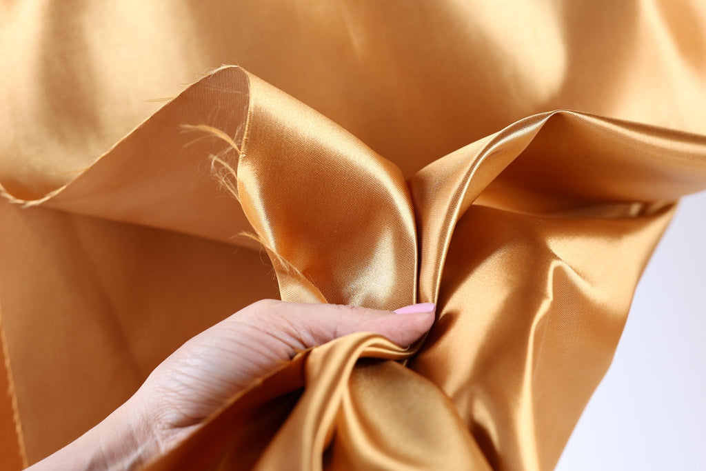 A hand is holding a piece of gold-colored fabric, which appears to be a shiny, smooth material.