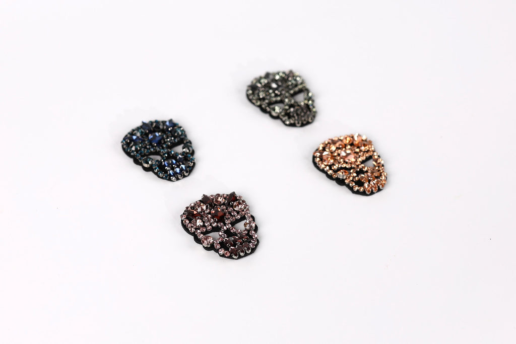 Four rhinestone-encrusted skull-shaped buttons in various colors, including black, blue, brown, and orange, arranged on a white background.