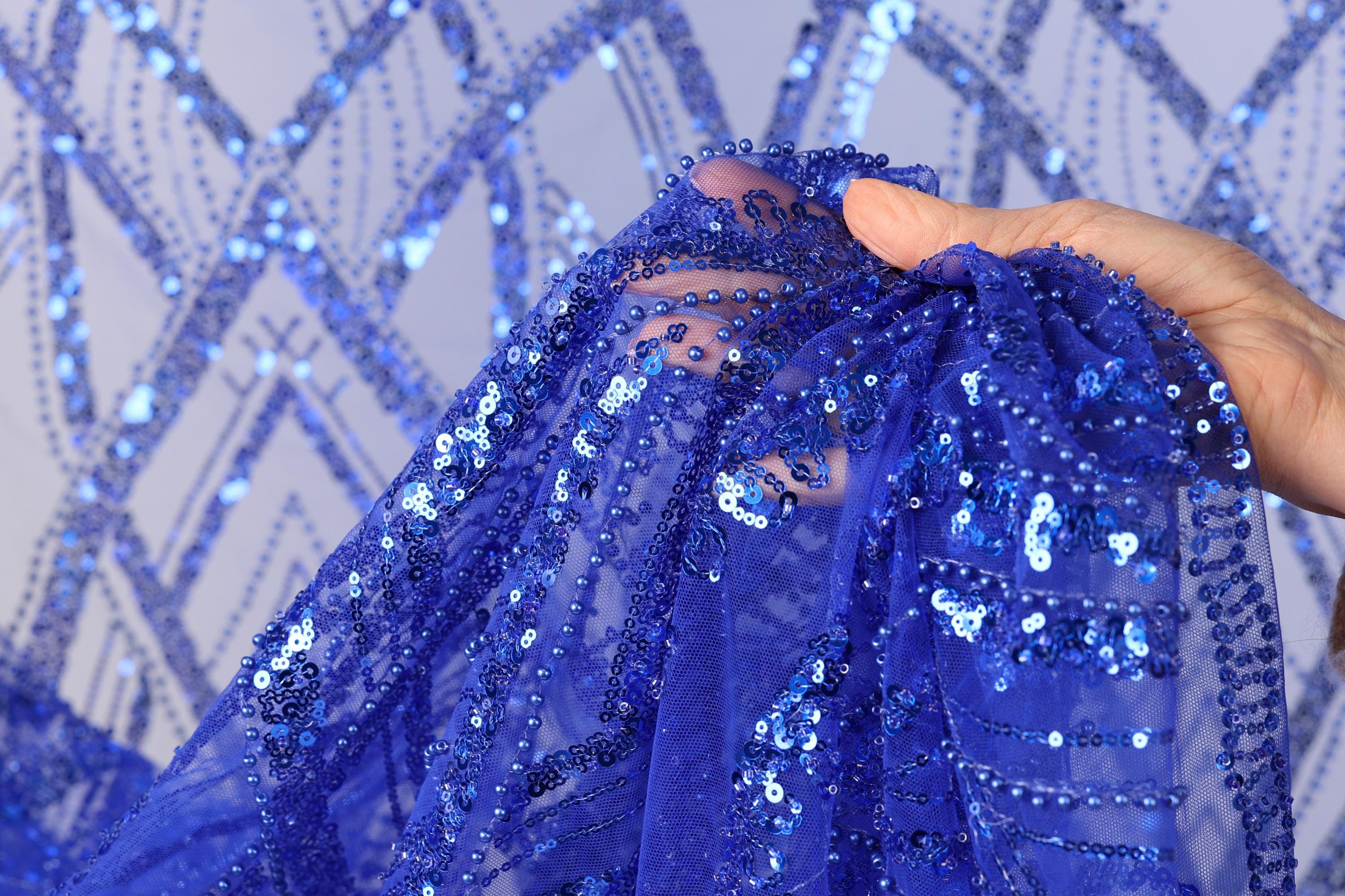A hand is holding a piece of blue sequin fabric, which appears to be a decorative textile.
