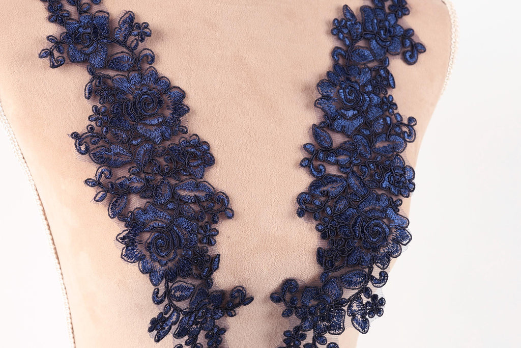 a close-up view of a mannequin torso wearing a dress with a floral lace overlay. The lace is intricately designed and appears to be made of a dark blue material, creating a striking contrast against the light-colored fabric of the dress.