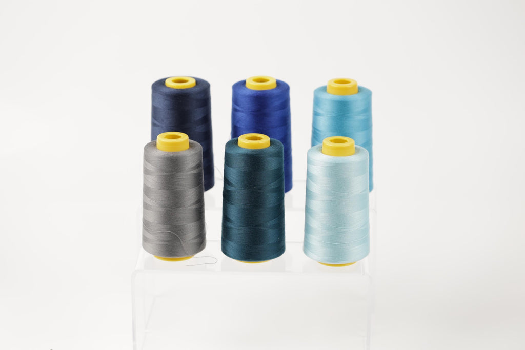 six spools of thread in various colors, including blue, gray, and turquoise, arranged in a neat row on a white background.