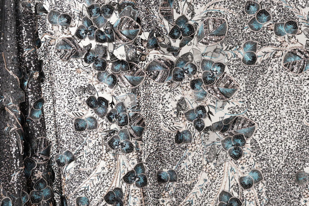 a close-up view of a fabric with a floral pattern. The fabric appears to be a blend of black and white, with accents of blue and green. The pattern consists of various shapes and sizes of flowers and leaves, creating a visually striking and intricate design. The fabric appears to be made of a shiny material, which adds to the overall visual appeal of the image.