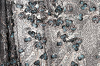 a close-up view of a fabric with a floral pattern. The fabric appears to be a blend of black and white, with accents of blue and green. The pattern consists of various shapes and sizes of flowers and leaves, creating a visually striking and intricate design. The fabric appears to be made of a shiny material, which adds to the overall visual appeal of the image.