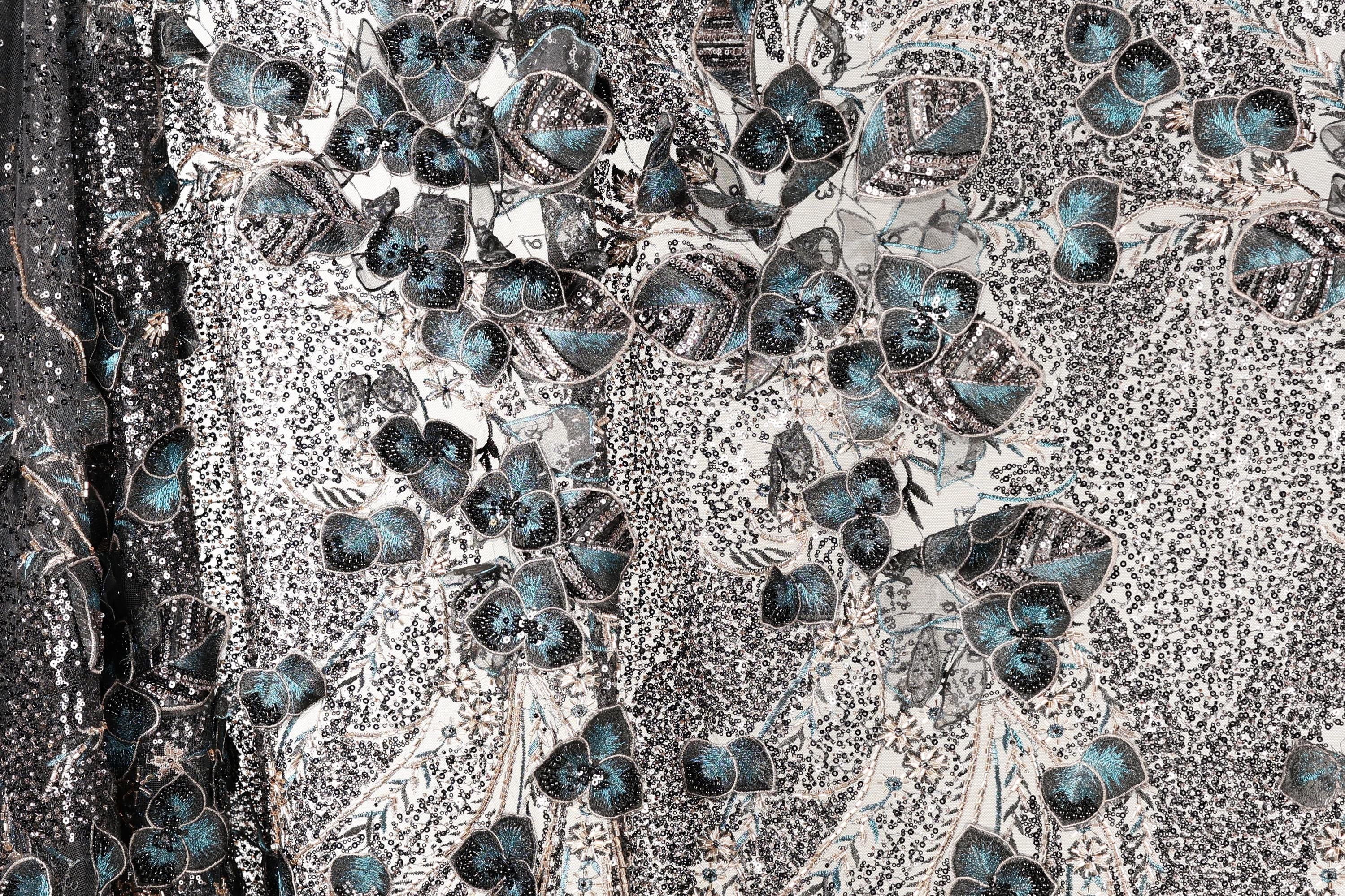 a close-up view of a fabric with a floral pattern. The fabric appears to be a blend of black and white, with accents of blue and green. The pattern consists of various shapes and sizes of flowers and leaves, creating a visually striking and intricate design. The fabric appears to be made of a shiny material, which adds to the overall visual appeal of the image.