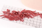 a close-up view of a decorative red lace or fabric with intricate floral and leaf-like patterns. The lace appears to be handmade and is placed on a ruler for scale.