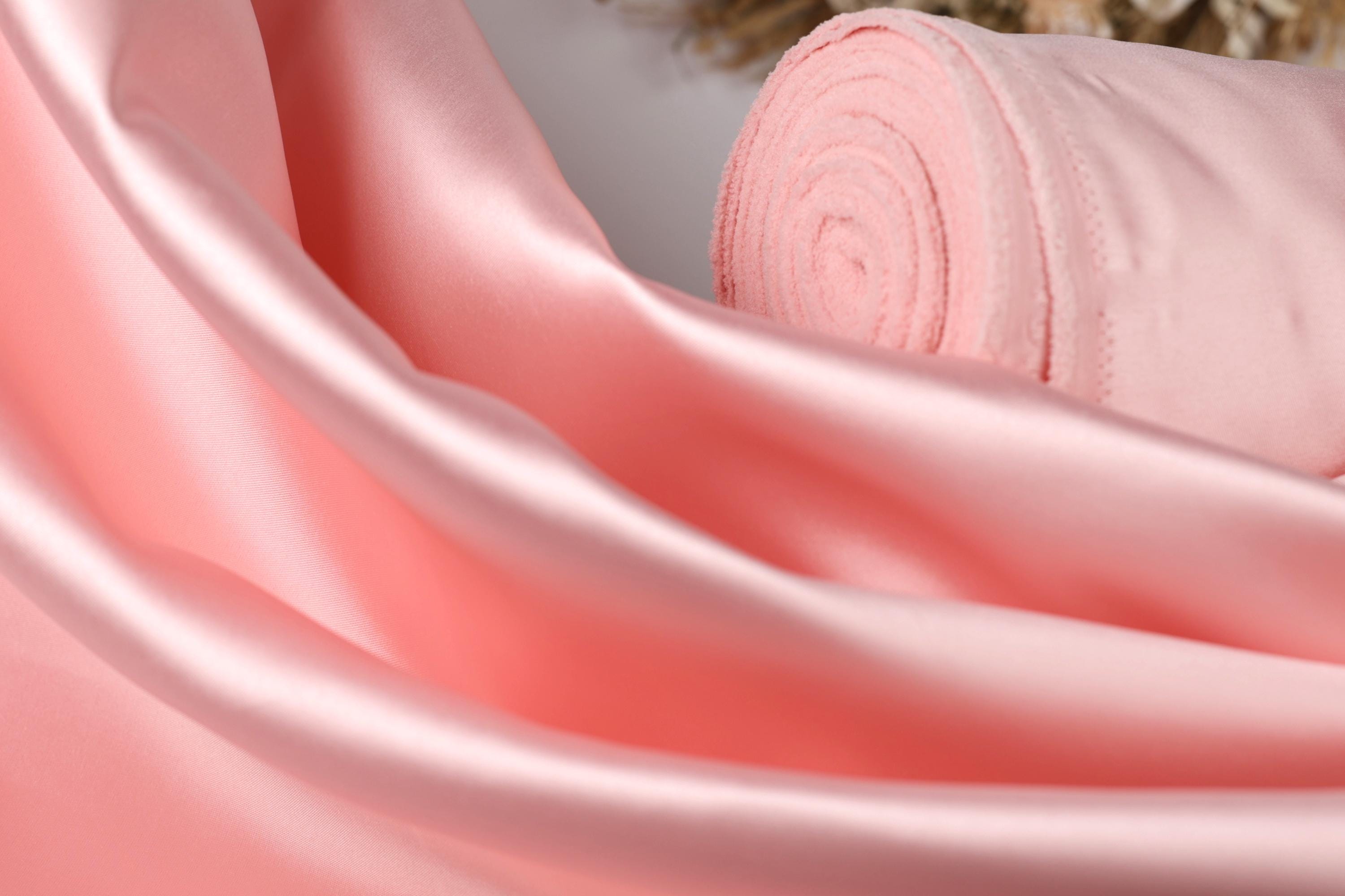 A close-up view of a soft, pink satin fabric, with a rolled-up piece of the fabric visible in the background.