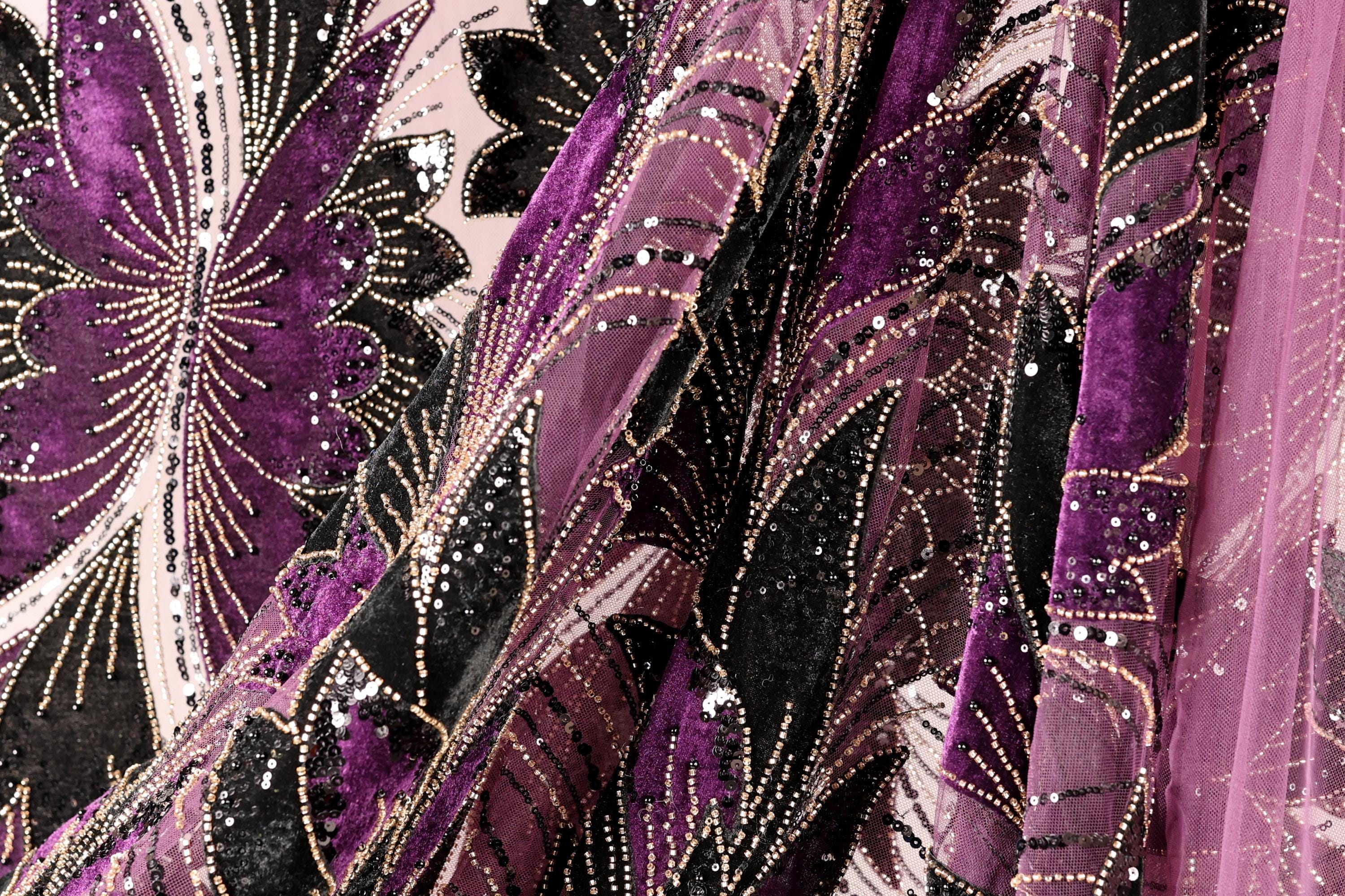 The image is a close-up of a fabric with a floral pattern in shades of purple and black, adorned with gold sequins.