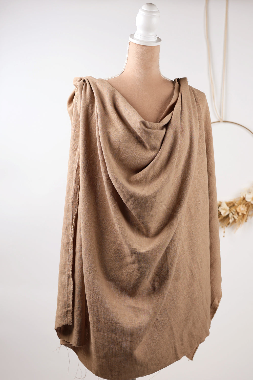 A mannequin wearing a beige, draped garment with a draped collar and sleeves, displayed against a white background.