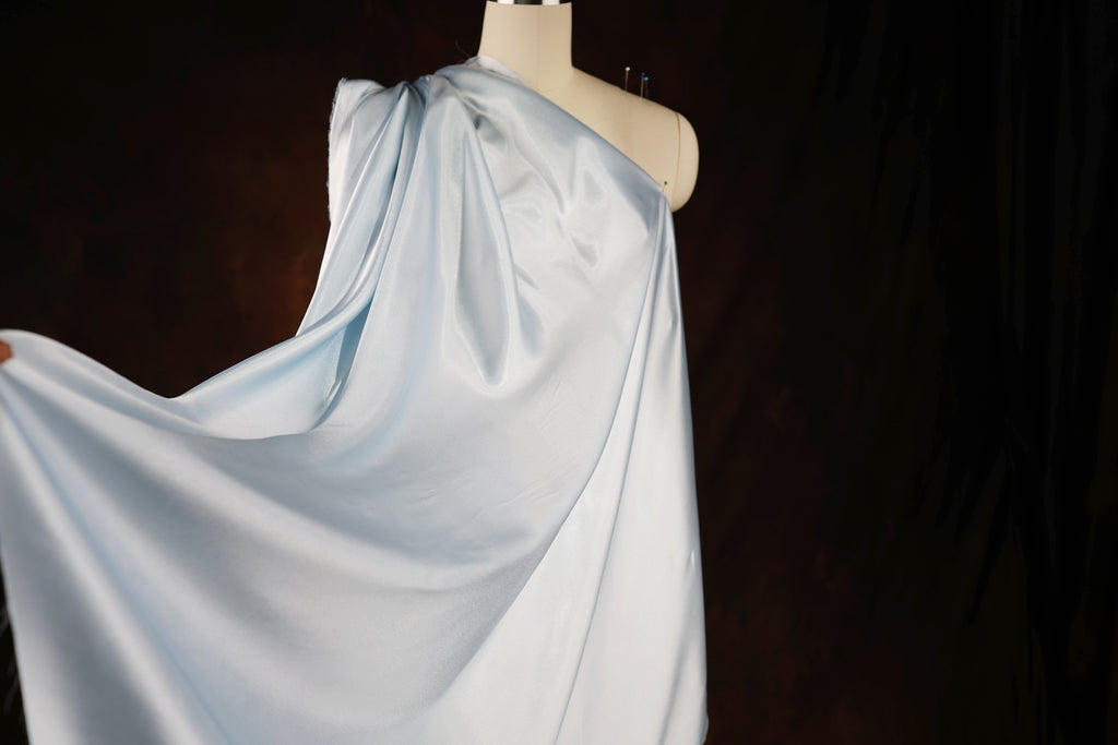 A mannequin wearing a flowing, light blue satin garment is displayed against a dark background.