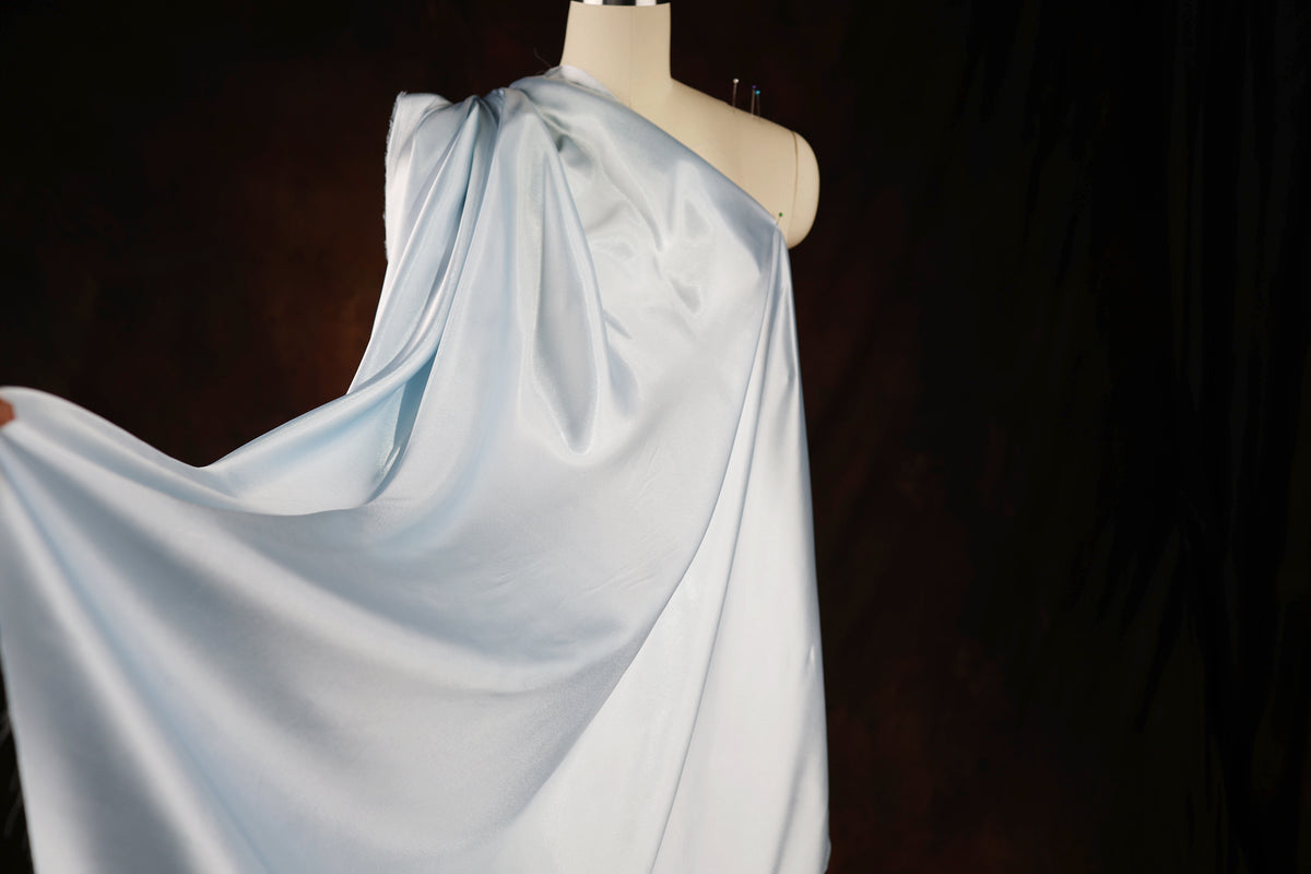A mannequin wearing a flowing, light blue satin garment is displayed against a dark background.