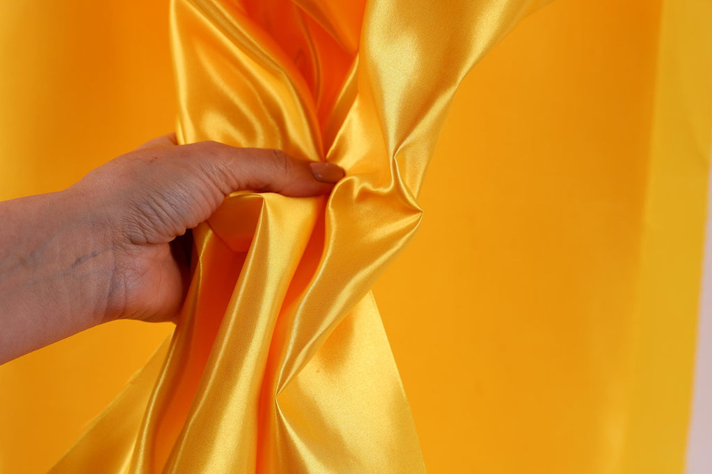 A hand is holding a yellow fabric, possibly silk or satin, against a bright yellow background.