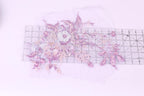 a close-up view of a delicate floral embroidery design, with various shades of pink and purple flowers and leaves. The design is displayed on a transparent surface, allowing for a clear view of the intricate details.