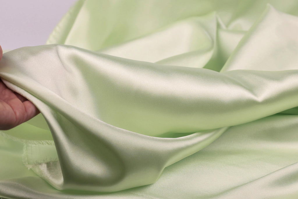A hand is holding a piece of light green satin fabric, which appears to be soft and smooth.