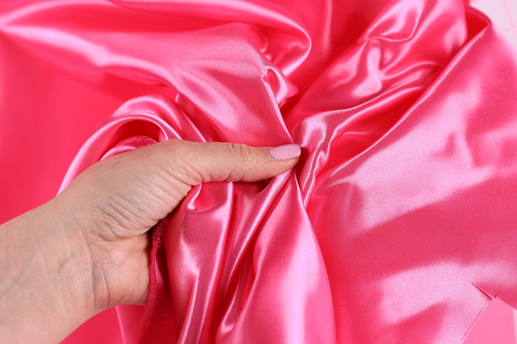 A hand is holding a piece of pink satin fabric, with the fingers gently touching the fabric.