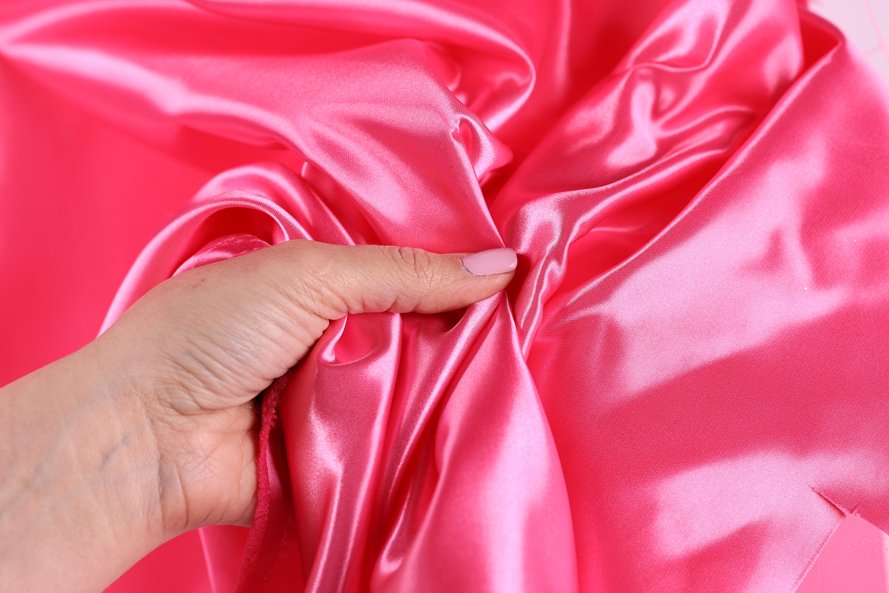 A hand is holding a piece of pink satin fabric, with the fingers gently touching the fabric.