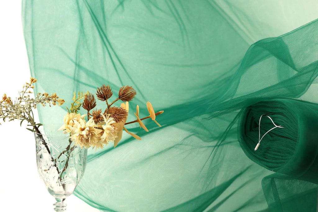a glass vase with flowers, a green fabric, and a white flower, all set against a green background.