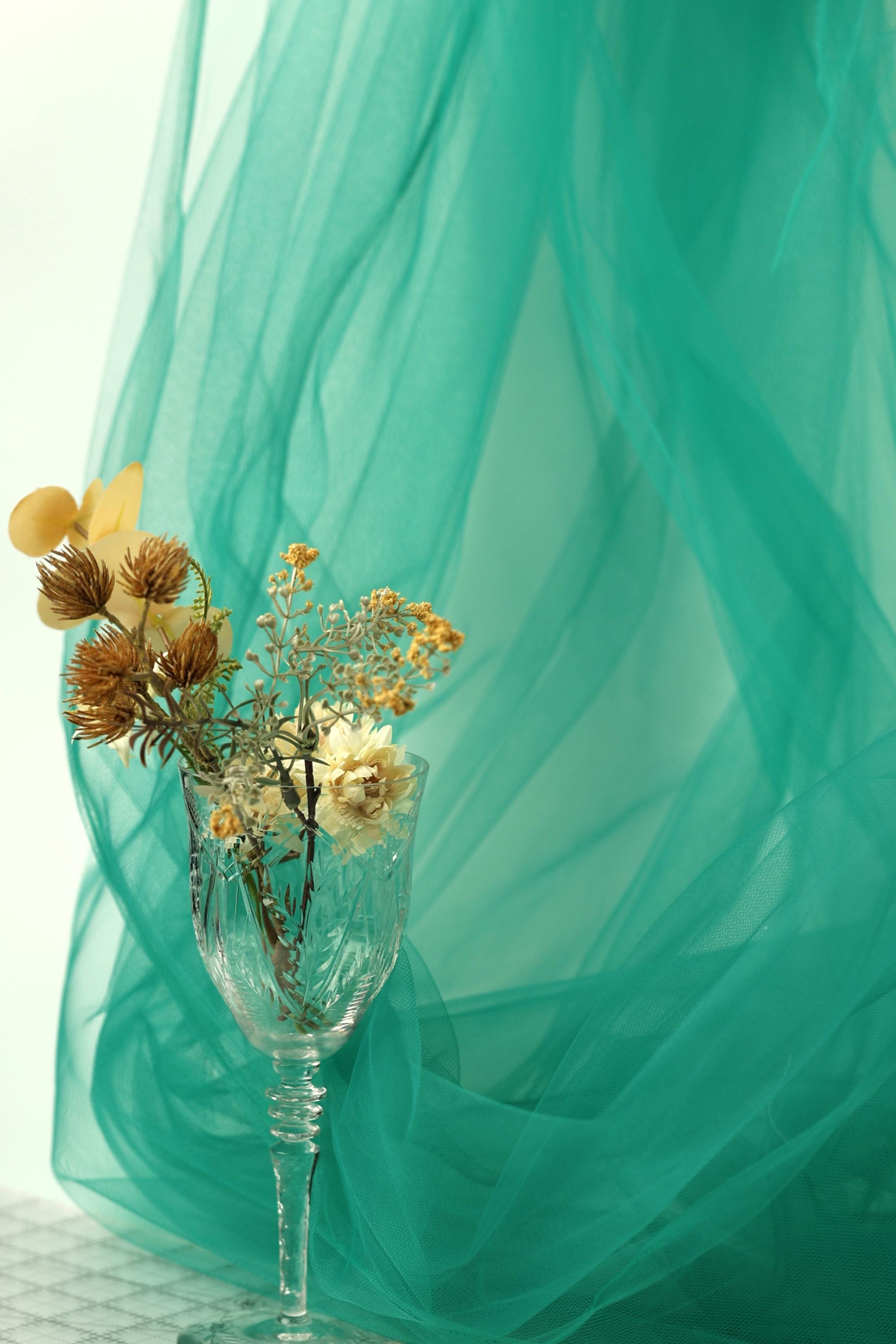 A glass vase filled with dried flowers sits on a table, with a green tulle curtain in the background.