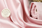 a pink satin fabric with a sewing artisan logo and a rolled-up pink fabric next to it.