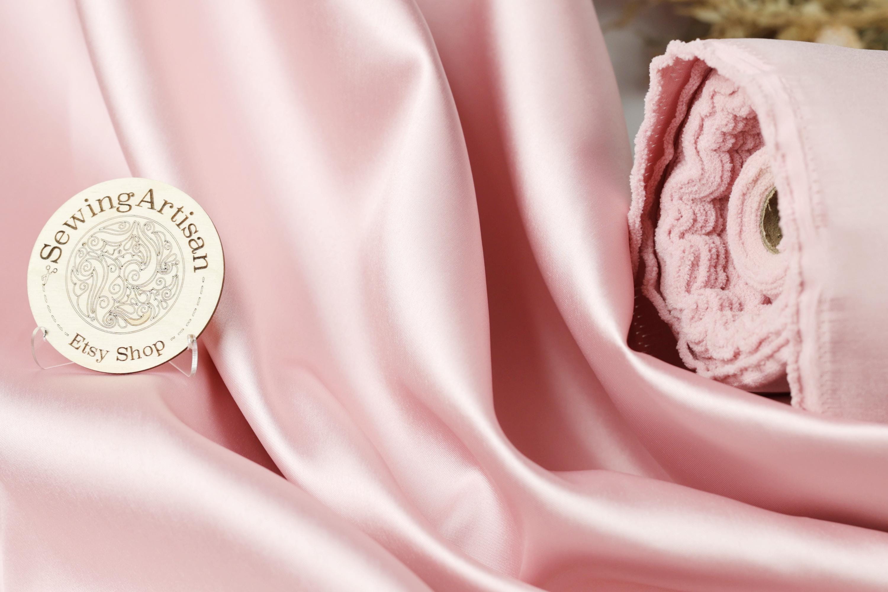 a pink satin fabric with a sewing artisan logo and a rolled-up pink fabric next to it.