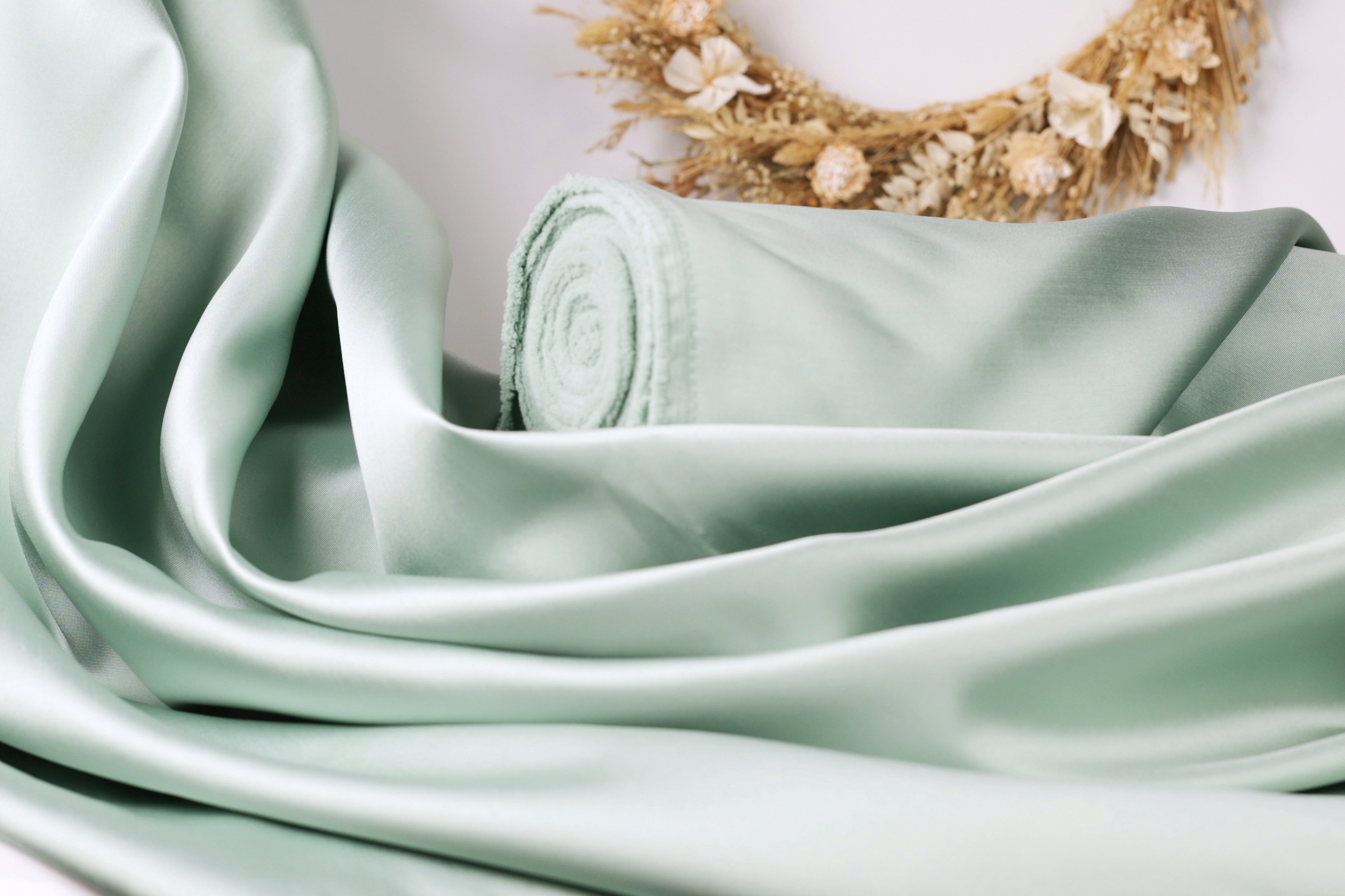 A close-up view of a light green satin fabric, possibly a sheer or satin material, with a wreath of dried flowers in the background.