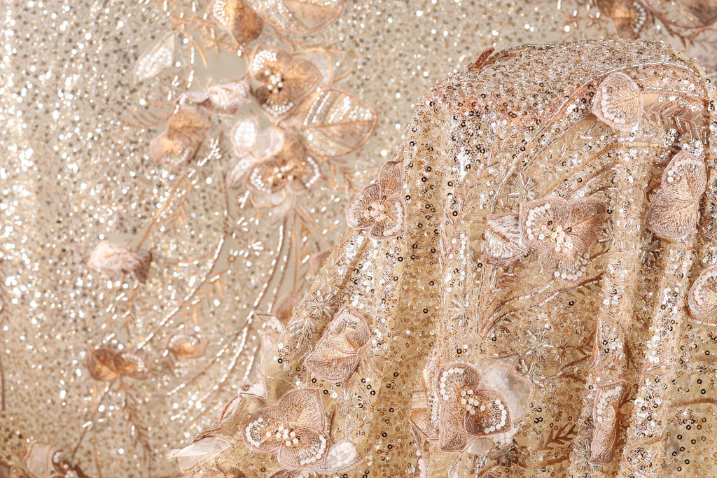 The image is a close-up of a fabric with a gold and beige color scheme, adorned with intricate floral and sequin designs. The fabric appears to be a sheer or sheer-like material, with a delicate and elegant appearance.