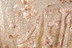 The image is a close-up of a fabric with a gold and beige color scheme, adorned with intricate floral and sequin designs. The fabric appears to be a sheer or sheer-like material, with a delicate and elegant appearance.