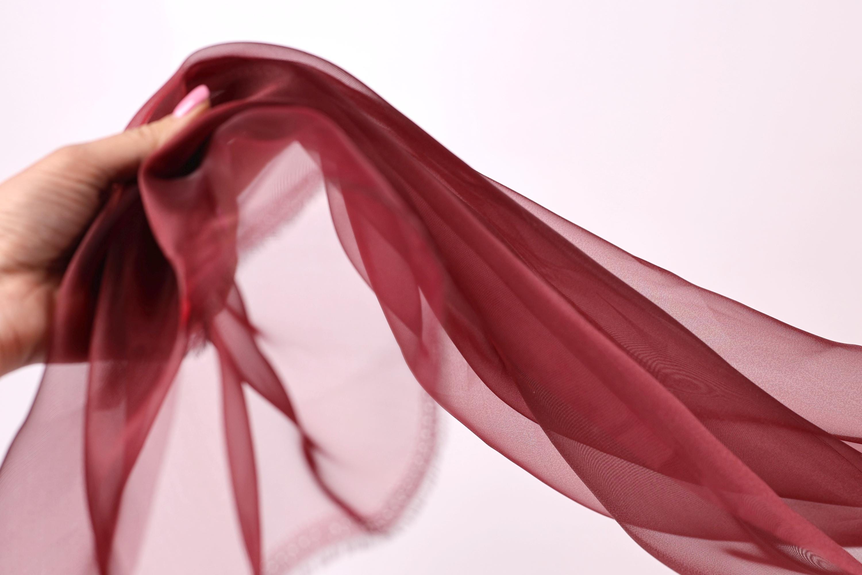 A hand is holding a long, flowing red fabric, which appears to be a scarf or shawl. The fabric is draped over the hand, creating a sense of movement and elegance.