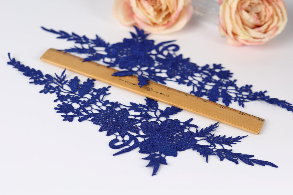 a close-up view of a blue lace fabric with intricate floral patterns, placed on a white surface. A wooden ruler is positioned in the center of the fabric, likely used for measuring or cutting the lace. In the background, there are two pink roses, adding a touch of color to the scene.